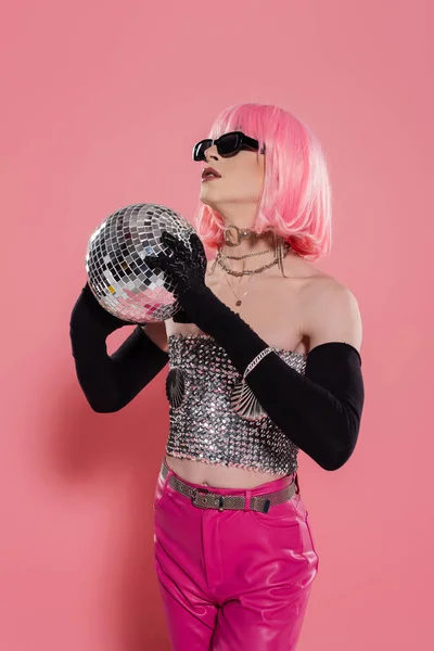 stock image Stylish transgender person in sunglasses and gloves holding disco ball on pink background 