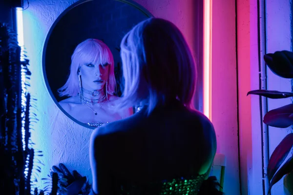 stock image Blurred transgender person in wig and top looking at mirror near plants and neon light at home 