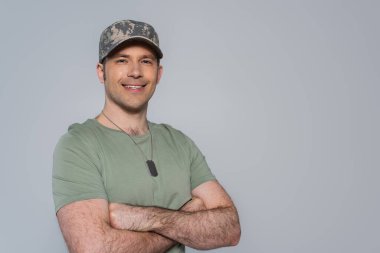 Mutlu Amerikan askeri tişörtlü ve askeri şapkalı Anma Günü boyunca kollarını kavuşturmuş gri renkte.