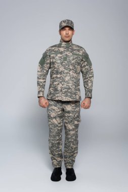 full length of patriotic soldier in military uniform and cap standing with clenched fists on grey  clipart