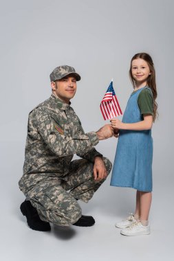 Neşeli küçük kız, gri gün anma gününde askeri üniformalı mutlu babaya Amerika bayrağı veriyor. 