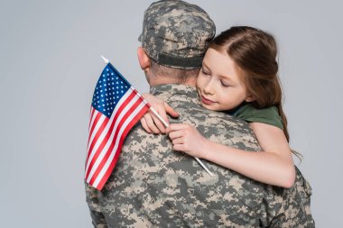 Askeri üniformalı bir asker, gri üzerinde Amerikan bayrağıyla kızına sarılıyor. 