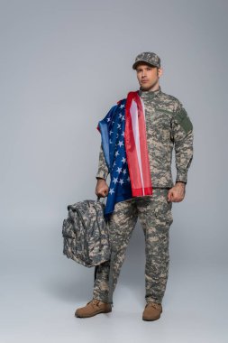 army soldier in camouflage uniform wrapped in flag of United States of America standing with backpack on grey  clipart