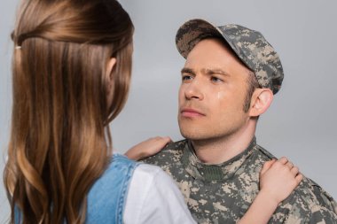 sad serviceman in military uniform crying and looking at daughter during memorial day isolated on grey  clipart