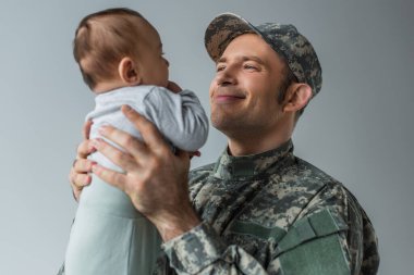 pleased army soldier in uniform holding newborn son in arms isolated on grey  clipart