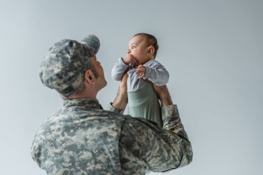 army soldier in uniform and cap holding baby boy in arms isolated on grey  clipart