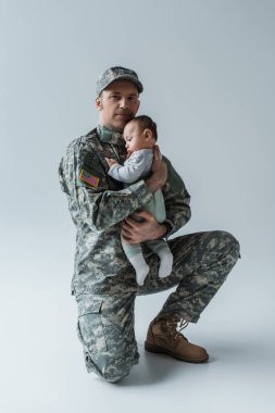 US soldier in uniform holding in arms newborn boy and looking at camera on grey clipart