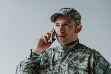 Başlık ve ordu üniformalı bir asker gri renkte izole edilmiş akıllı telefondan konuşuyor. 