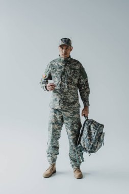 Amerikan askeri sırt çantası tutarken akıllı telefon kullanıyor ve gri üzerinde duruyor. 