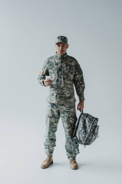 American soldier using smartphone while holding backpack and standing on grey  clipart