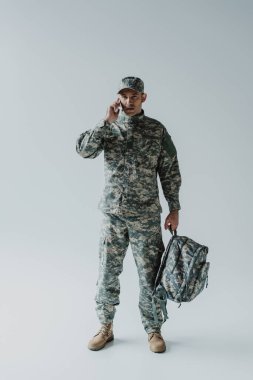 Asker üniformalı bir asker, gri bir çantayla akıllı telefondan konuşuyor. 