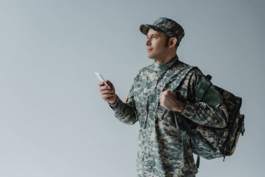 Asker üniformalı, şapkalı bir asker sırt çantalı ve akıllı telefonu gri renkte tutuyor. 