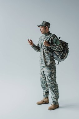 military man in cap and army uniform standing with backpack and holding smartphone on grey  clipart