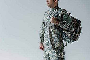 Asker üniformalı askerin gri üzerinde sırt çantasıyla dikilirken çekilmiş görüntüsü. 