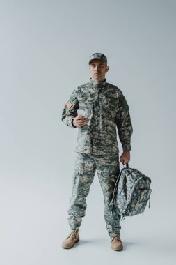 American soldier in army uniform with national flag holding smartphone and backpack on grey 
