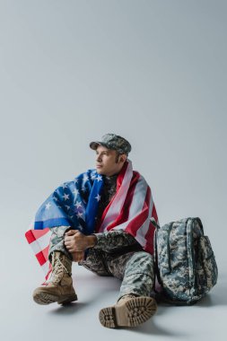 Amerikan askeri üniformalı ve ABD bayrağı gri sırt çantasının yanında oturuyor. 