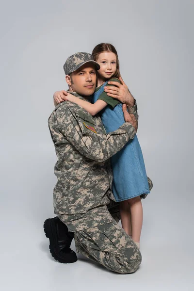 preteen girl hugging father in military uniform and cap during memorial day on grey