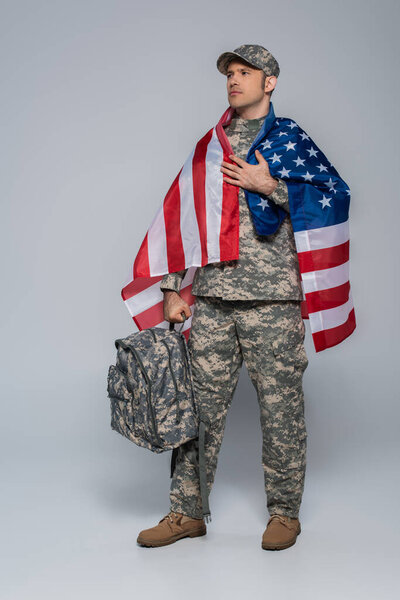 patriotic army soldier in camouflage uniform wrapped in flag of United States of America standing with backpack on grey 