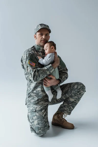 US soldier in uniform holding in arms newborn boy and looking at camera on grey