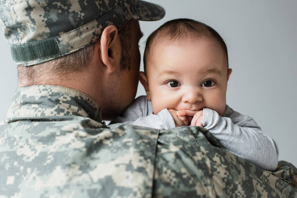 man in army uniform and cap hugging newborn son isolated on grey 