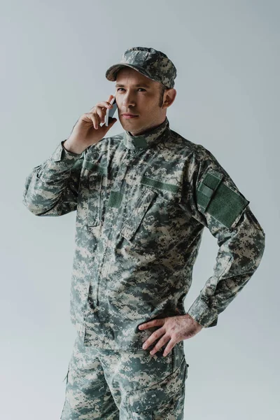 キャップと軍の制服を着た軍人が灰色で隔離されたスマートフォンで話しています — ストック写真