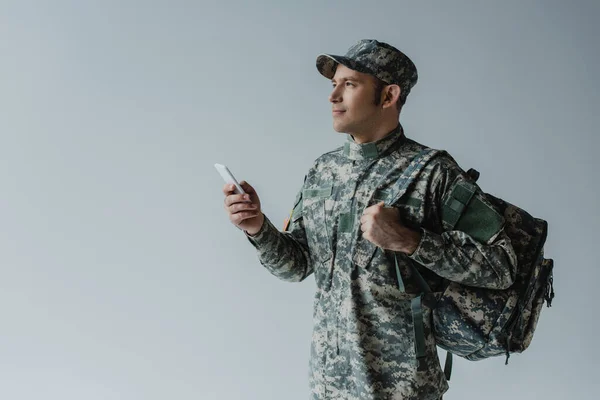 キャップと軍の制服を着た軍人がリュックを持ってグレーに隔離されたスマートフォンを持ち — ストック写真
