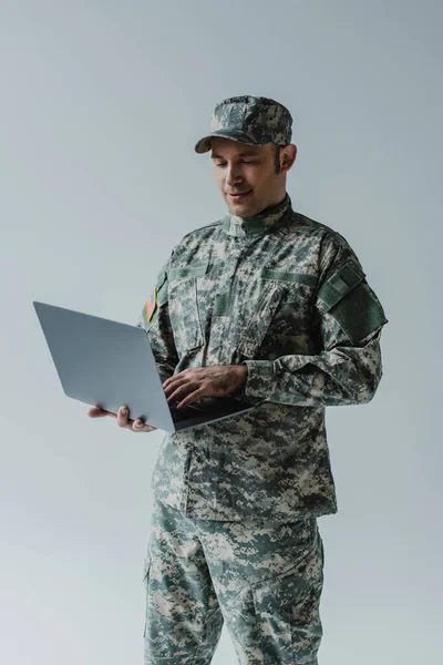 グレーに隔離されたノートパソコンを使って軍の制服と帽子を着たいい兵士だ — ストック写真