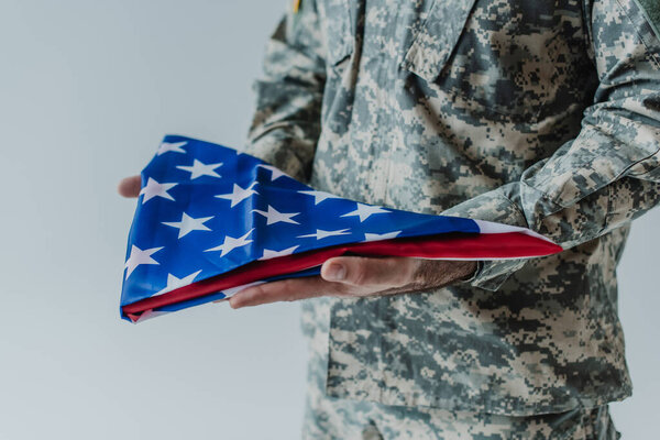 partial view of American soldier in uniform holding folded flag of United States during memorial day isolated on grey 