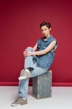 Koyu kırmızı arka planda oturan kot elbiseli genç bir model.