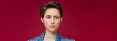 suspicious young woman with short hair looking away on burgundy background, banner  clipart