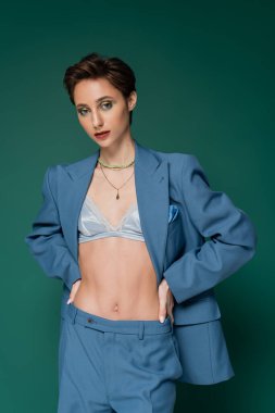 young woman in blue suit with silk bra underneath posing with hands on hips on turquoise green background  clipart