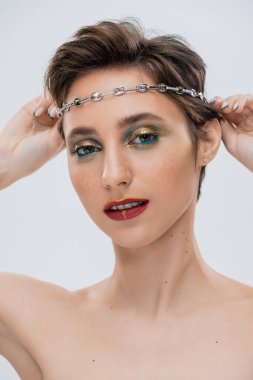 young woman with shiny makeup and short hair wearing silver chain on head isolated on grey  clipart
