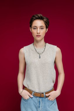 young woman in sleeveless shirt and chain necklace standing on maroon background  clipart