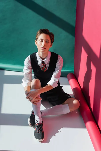 stock image full length of stylish young woman with short hair sitting in school uniform on pink and green 