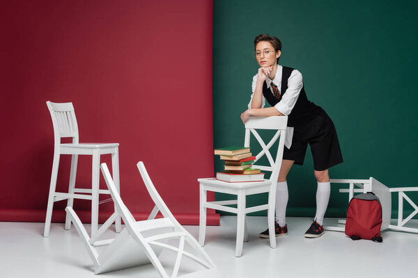 полная длина стильный молодой студент с короткими волосами опираясь на стул с книгами на зеленом и розовом фоне 