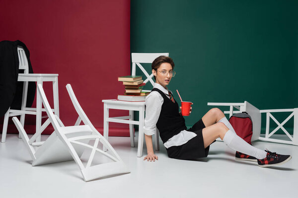 стильный молодой студент в очках, держа напиток в пластиковой чашке возле стульев и книг на зеленом и бургундском фоне 