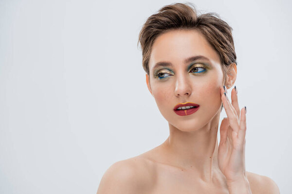 young woman with shimmery eye makeup and short hair touching cheek isolated on grey 
