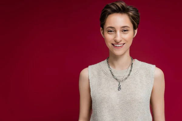 pleased young woman in sleeveless shirt and chain necklace standing on maroon background