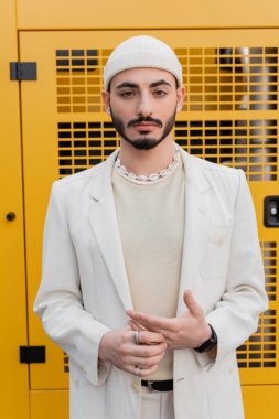 Şapkalı ve ceketli, dışarıda kameraya bakan şık bir gay portresi. 