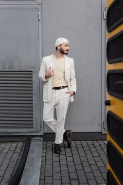 Şık giyinmiş, şapkalı ve takım elbiseli bir gay. Şehir sokağının yakınında poz veriyor. 