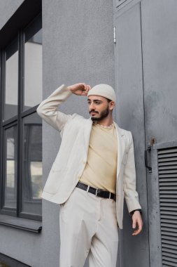 Bej takım elbiseli ve şapkalı modaya uygun homoseksüel bir adam binanın yakınına bakıyor. 