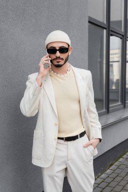 Güneş gözlüklü ve bej takım elbiseli modacı açık havada akıllı telefondan konuşuyor.