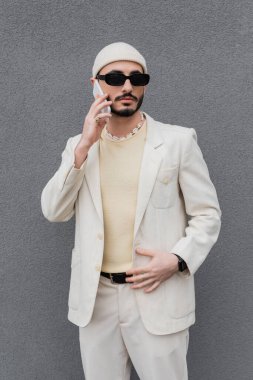 Takım elbiseli ve güneş gözlüklü modacı açık hava binasının yakınında akıllı telefondan konuşuyor. 