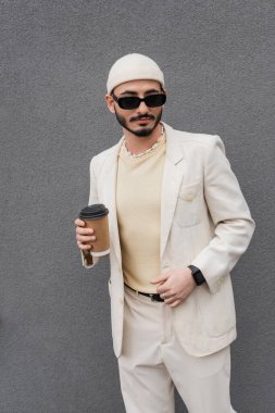 Bej takım elbiseli, güneş gözlüklü, dışarıda kağıt bardak tutan modaya uygun bir gay. 