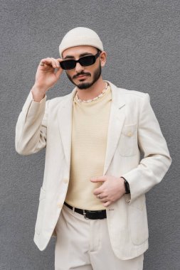 Açık havada güneş gözlüğüne dokunan bej takım elbiseli moda homoseksüel adam. 
