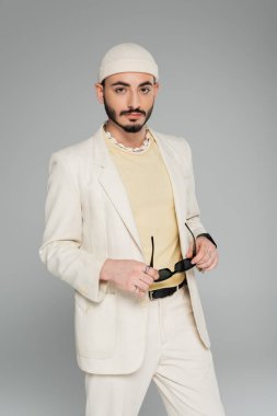 Takım elbiseli, şapkalı, güneş gözlüklü ve gri renkli kameraya bakan şık bir gay portresi.  