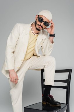 Güneş gözlüklü ve takım elbiseli moda eşcinsel adam gri sandalyenin yanında poz veriyor.  