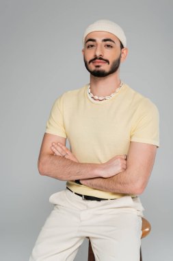 Gri renkli sandalyenin yanında kollarını kavuşturan şapkalı modaya uygun eşcinsel portresi.  