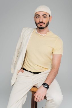 Gri renkli sandalyede oturan ceketli ve şapkalı, modaya uygun eşcinsel bir adamın portresi.  