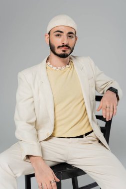 Sakallı, takım elbiseli, deniz kabuğu kolyeli eşcinsel adam gri sandalyede tek başına oturuyor.  